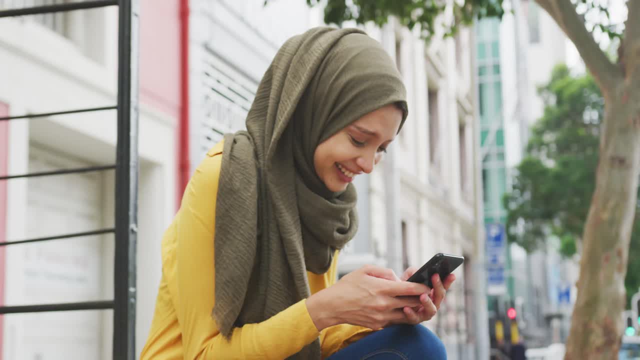 Woman wearing hijab using her phone in the street and laughing