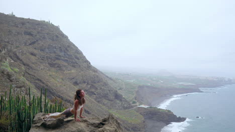 Amidst-island-landscapes,-the-woman-sits-on-a-cliff,-engaging-in-a-dog-pose,-inhaling-the-sea-air,-and-embracing-the-essence-of-yoga-by-the-ocean
