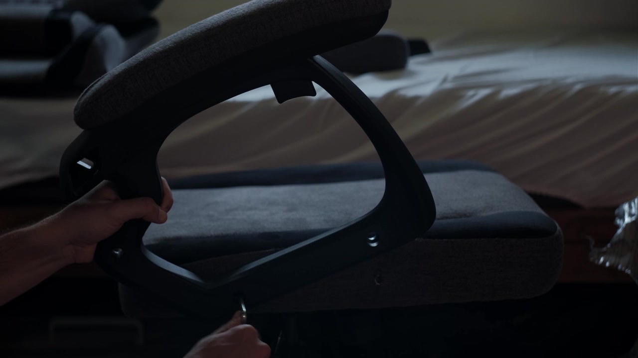 Plano medio estático de manos ajustando el reposabrazos de una silla gaming durante el montaje de muebles en un espacio de trabajo interior con poca luz