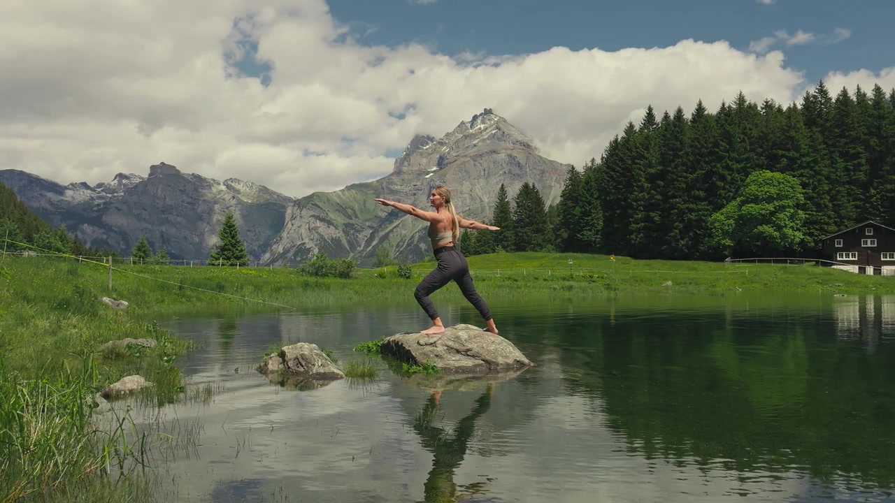 Donna caucasica pratica una posa yoga del guerriero su una roccia in un lago alpino incontaminato. Una drammatica cima montuosa si erge dietro di lei, creando una serena scena di benessere nelle Alpi svizzere.