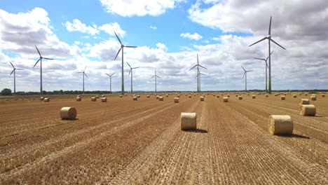 Ein-Atemberaubendes-Luftvideo-Fängt-Den-Anmutigen-Tanz-Der-Windkraftanlagen-Auf-Dem-Frisch-Abgeernteten-Feld-Eines-Bauern-Aus-Lincolnshire-Ein,-Mit-Dem-Zusätzlichen-Charme-Goldener-Heuballen