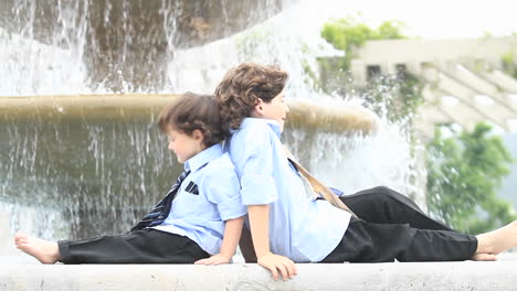 Ein-Paar-Jungen-In-Festlicher-Kleidung-Sitzen-Rücken-An-Rücken-Vor-Einem-Brunnen-1