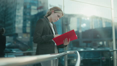 secretary reading red folder near iron railing outside glass office building reflecting cityscape with cars and sunlight ambiance, showcasing professionalism, focus, and corporate atmosphere