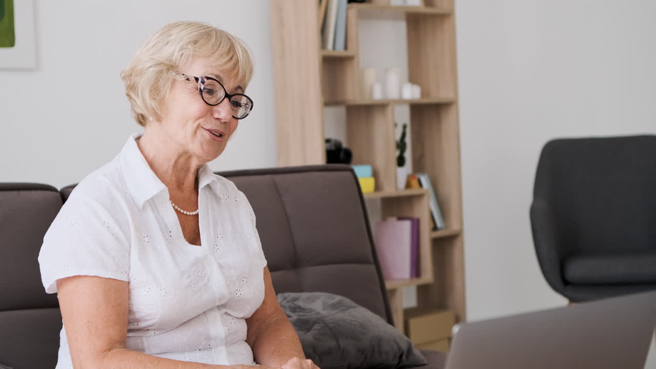 Smiling senior woman sitting on sofa in living room talking on video call