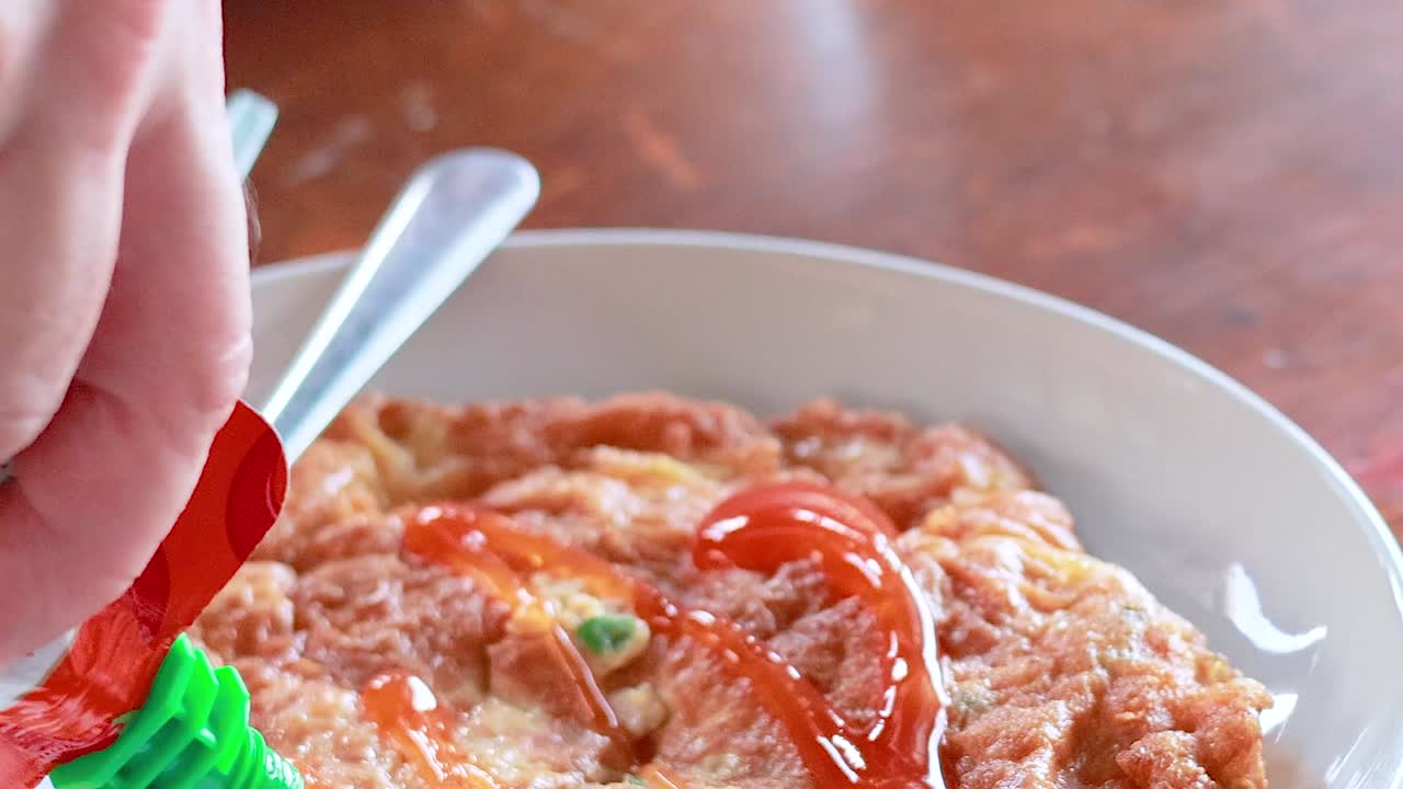 A hand drizzles sauce over an omelette, accompanied by a fork and spoon on a wooden table.