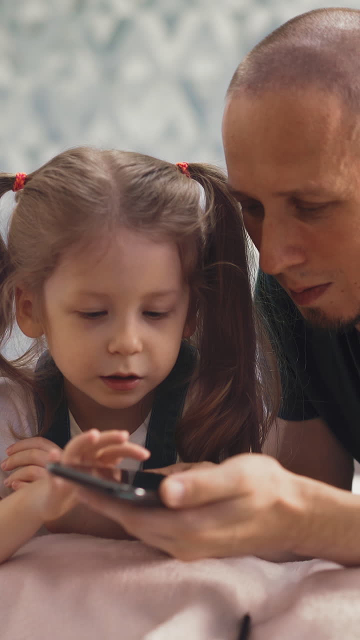 close up of young bald dad and little daughter with long hair, together set up online learning on smartphone, tap fingers, flip through, swipe
