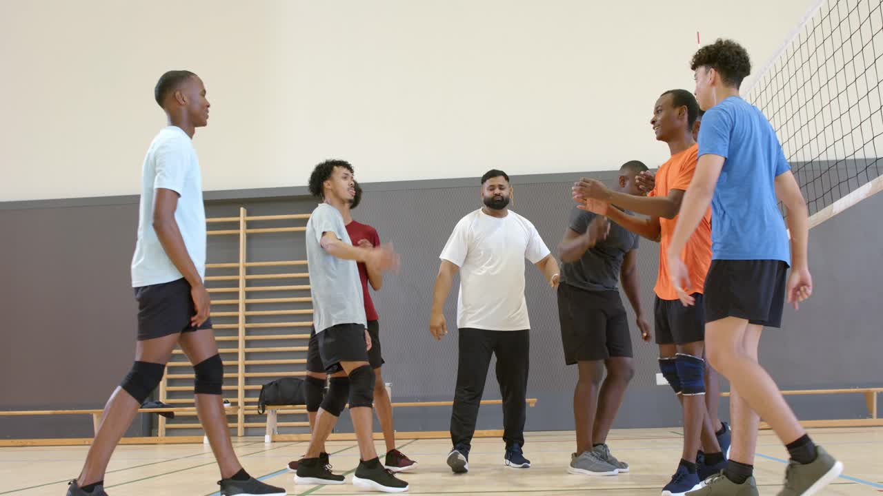 Vielfältiges Herren-Volleyballteam legt im Fitnessstudio am Netz die Hände übereinander und skandiert Parolen, nachdem der Trainer einen Anfeuerungsruf heruntergezählt hat.