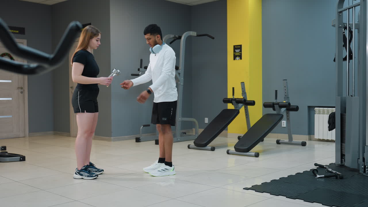 young man in white top handing over dumbbell to woman in black sportswear as she prepares for stamina-building exercise during indoor gym session with various fitness equipment visible in background