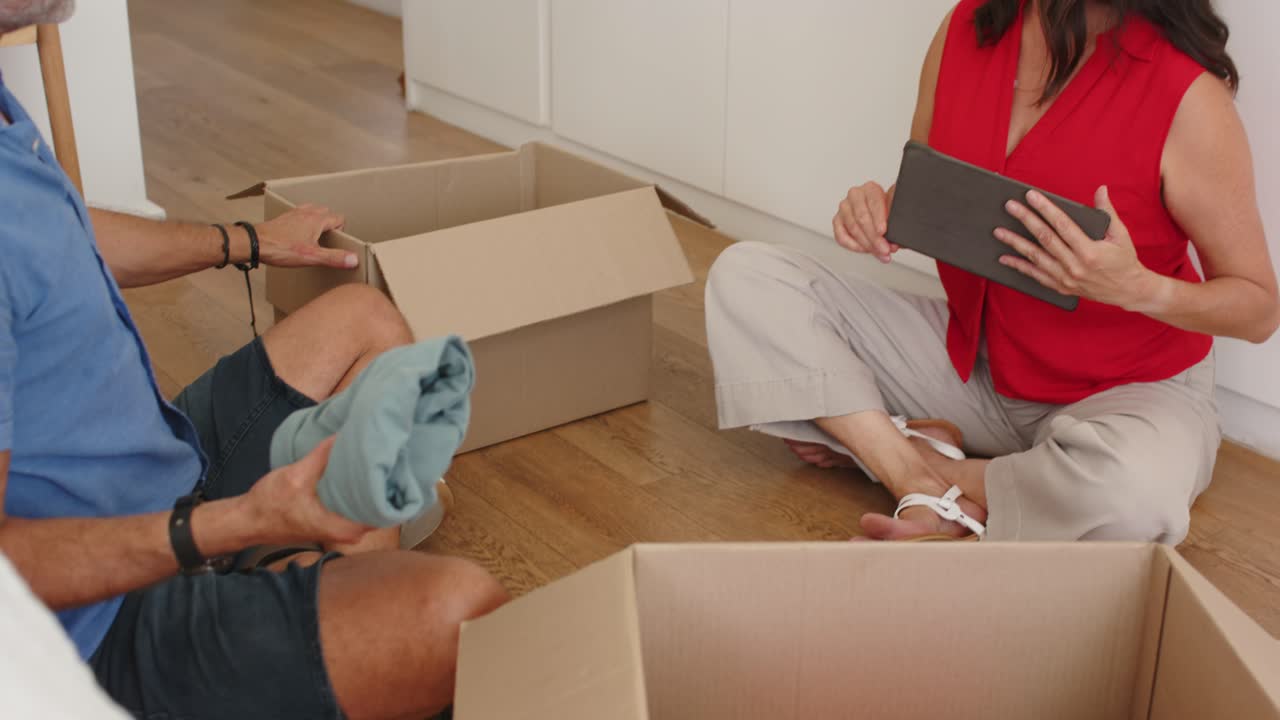 Mature couple sitting on floor holding tablet, man showing shirt placing into DONATION-labeled box