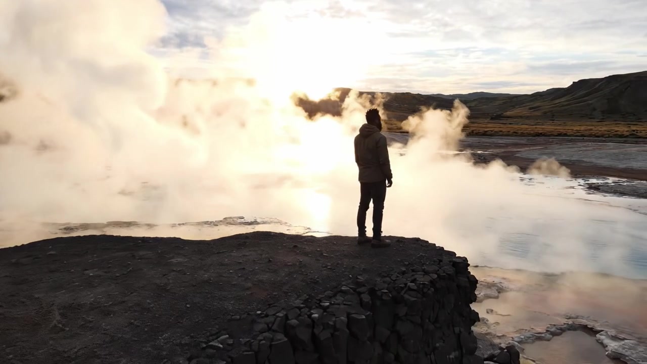 Un momento sereno: Una figura in silhouette si erge ai margini di un geyser fumante, bagnata da una luce dorata mentre il sole tramonta su un paesaggio tranquillo.