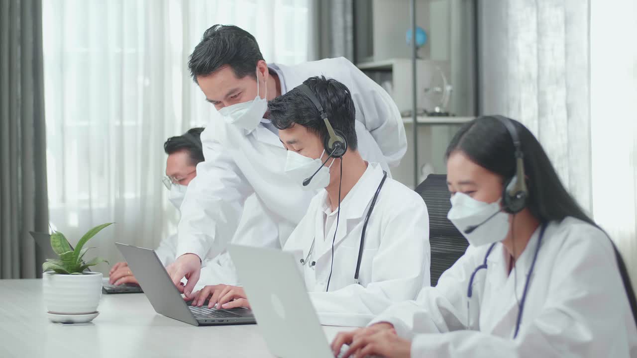A Man Coming To Help One Man Of Three Asian Doctors In Headsets And Masks Using Computer For Working As Call Center Agent While His Colleagues Are Speaking And Typing During A Call At The Office.