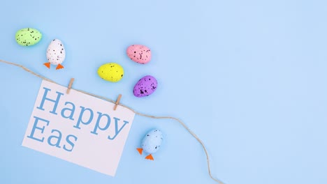 happy easter write on paper card note hooked on rope on pastel blue background with eggs. stop motion flat lay