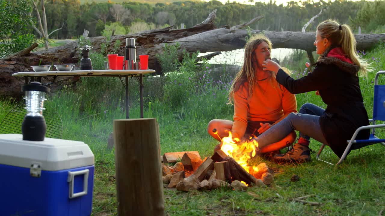 캠프 불 4k 근처에 마시멜로와 함께 남자를 먹이는 젊은 여성