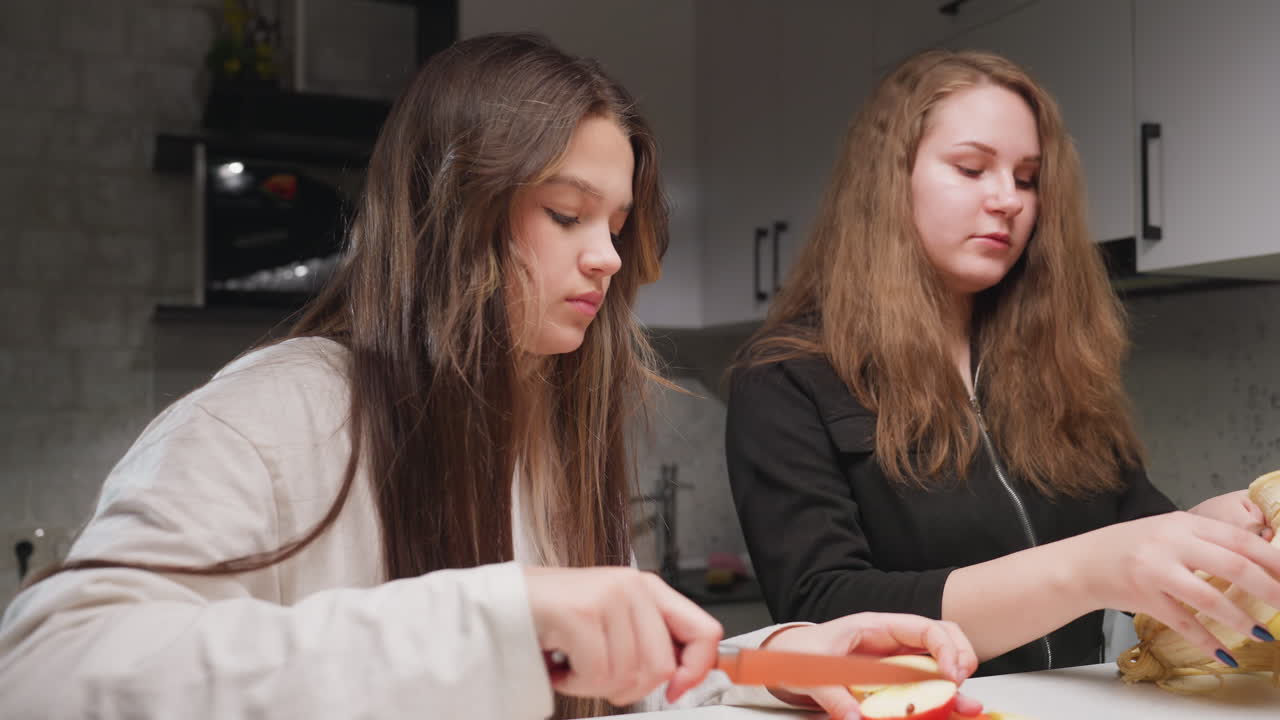 Students in kitchen at table, one uses chopping board and knife to cut red apple while partner peels banana, calm preparation scene, clean counter, soft indoor light, healthy snack moment