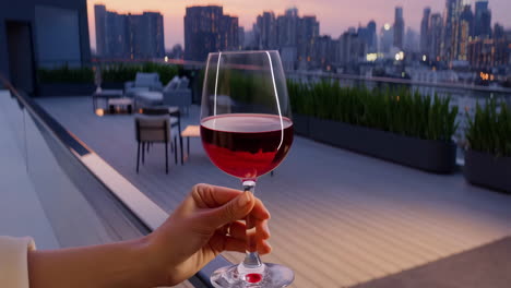 hand holding a glass of red wine on a rooftop at sunset with a city view