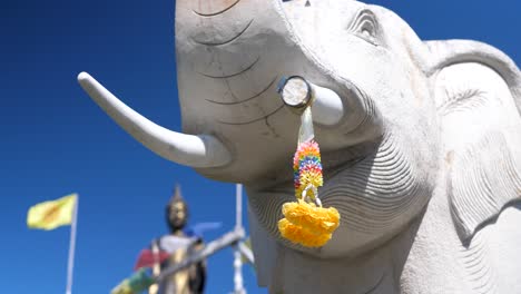 statue of ganesha, elephant, with flower offering on tusk, prayer flags and buddha statue in background