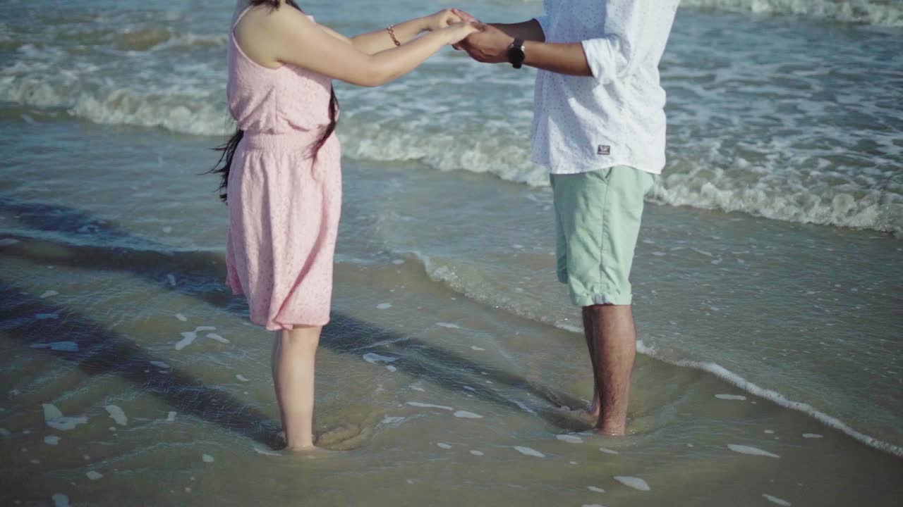 sin foto en la cara de una pareja tomándose de la mano y jugando cerca de las olas en una playa