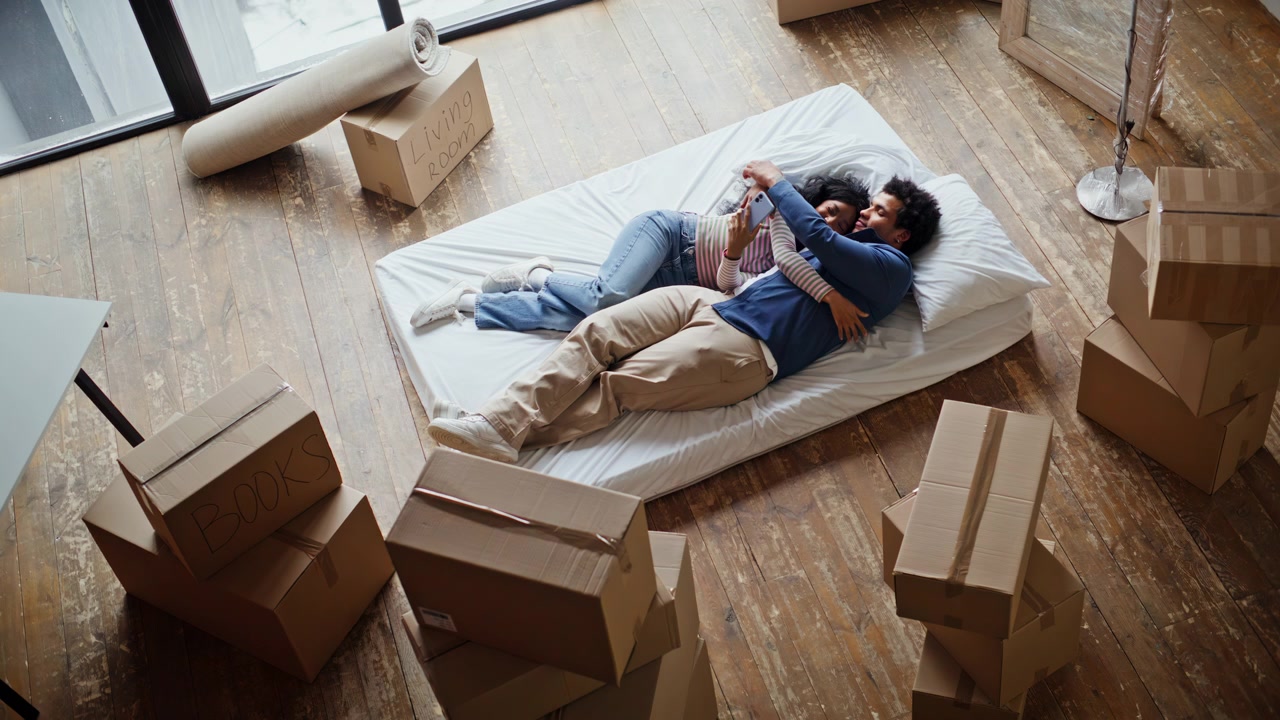 Casal deitado segurando o smartphone, abraçados no colchão, rindo no novo apartamento