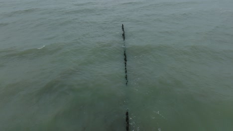 Luftaufnahme-Aus-Der-Vogelperspektive-Eines-Alten-Hölzernen-Piers-An-Der-Ostseeküste,-Bewölkter-Wintertag,-Weißer-Sandstrand,-Holzpfähle,-Wellen,-Die-Auf-Die-Küste-Treffen,-Breite-Drohnenaufnahme,-Die-Sich-Rückwärts-Tief-Bewegt