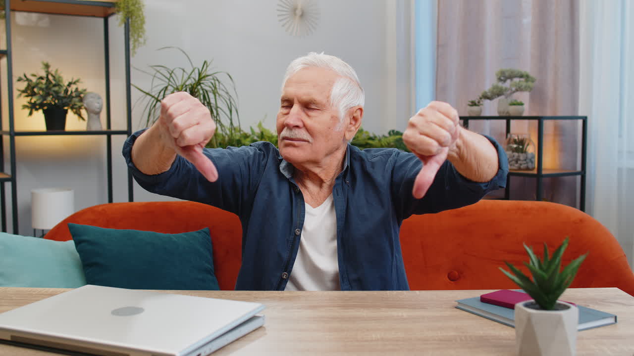 Upset senior man showing thumbs down dislike bad work disapproval dissatisfied feedback at home