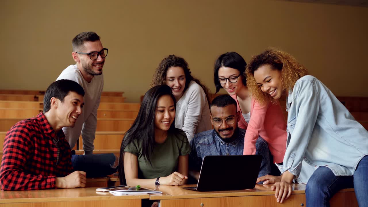 Cheerful men and women are working with laptop looking and pointing at screen, talking and laughing sitting at desks in university lecture hall. Students and technology concept.