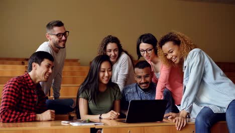 cheerful men and women are working with laptop looking and pointing at screen, talking and laughing sitting at desks in university lecture hall. students and technology concept.