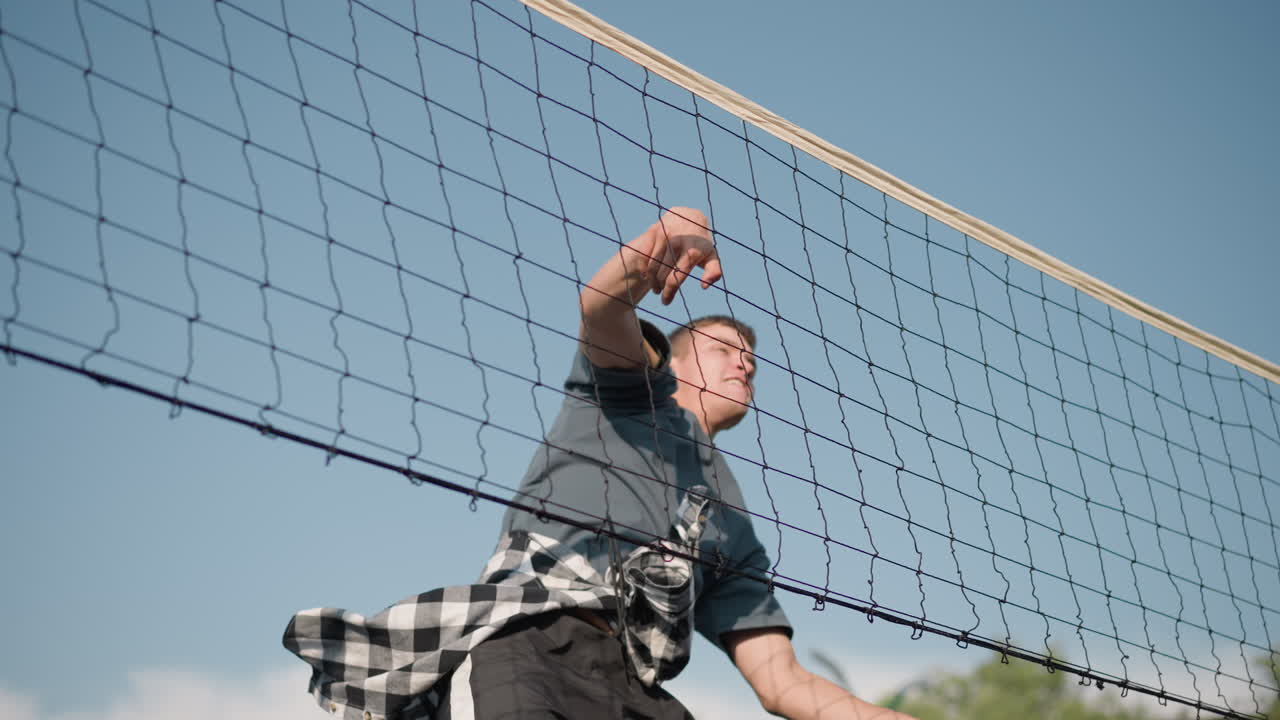 강렬한 훈련 기간 동안 배구 공을 다시 네트 위로 던지려는 남자가 높이 뛰어내립니다. 배경에는 다양한 장비가 있는 야외 스포츠 시설이 있습니다.