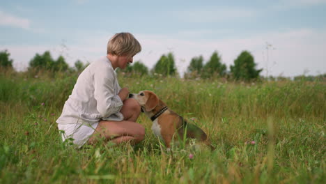 dog owner interacting playfully with her pet dog as they share a tender moment in grassy field, owner is squatting down while dog gazes up and appears to be taking something from her hand