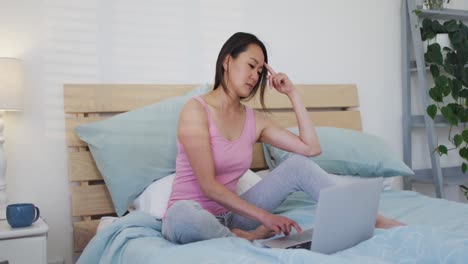 Happy-asian-woman-sitting-on-bed,-using-laptop-in-the-morning