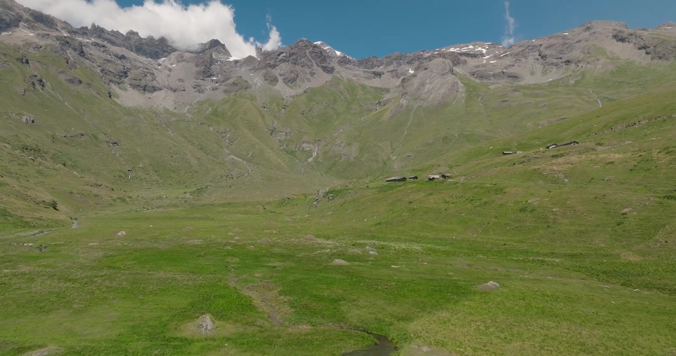 Un drone sorvola la parete di una diga e segue un bacino alpino turchese verso la sua sorgente montana, rivelando cime aspre, acqua incontaminata e uno spettacolare paesaggio d'alta quota