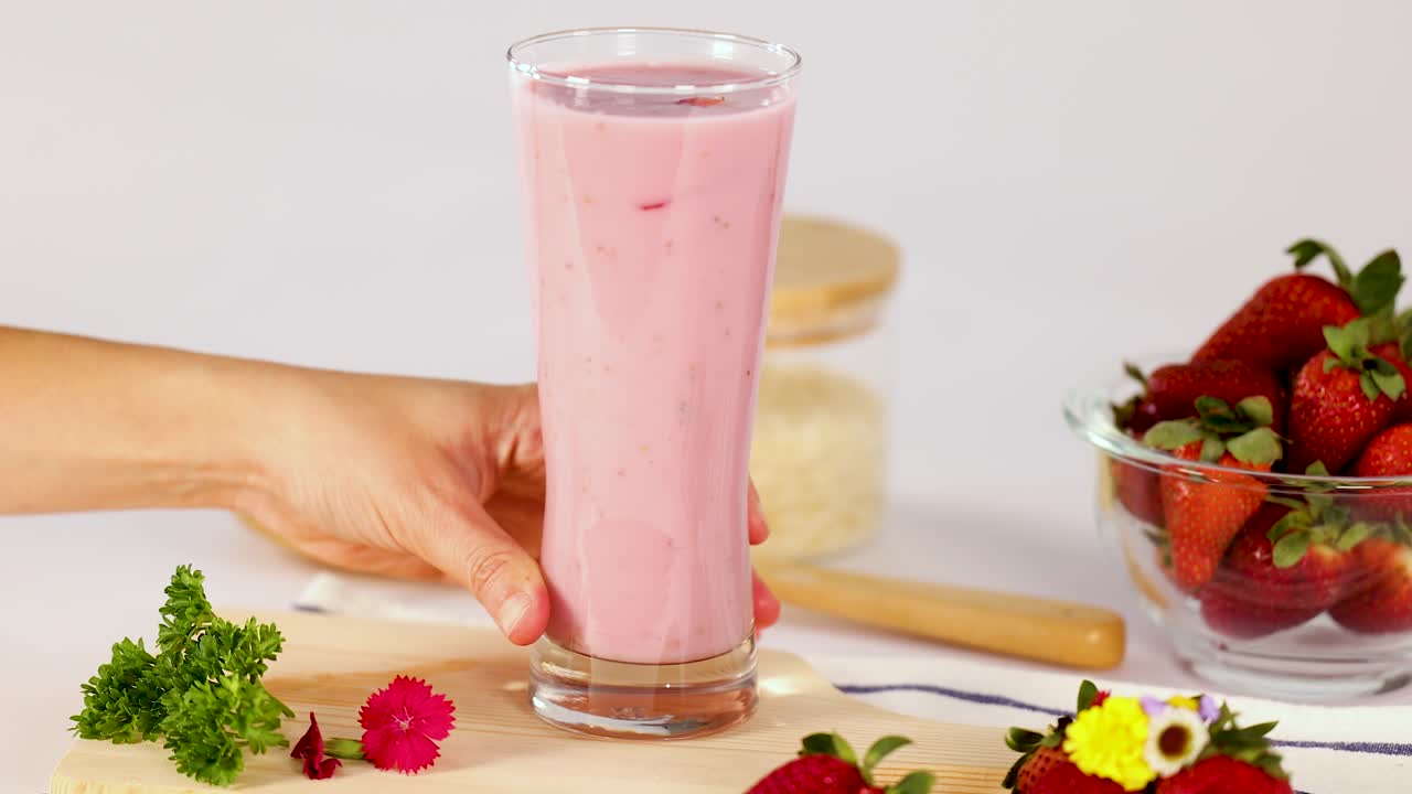 Una mano coloca un vaso alto de batido de fresa en una mesa de cocina luminosa, con luz natural