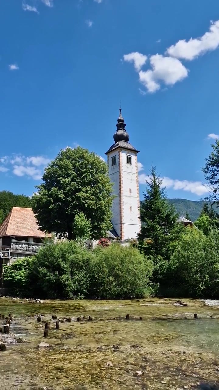 Vista verticale del campanile di una chiesa cattolica vicino al fiume, emissario del Lago di Bohinj, Slovenia, in una calda giornata di sole