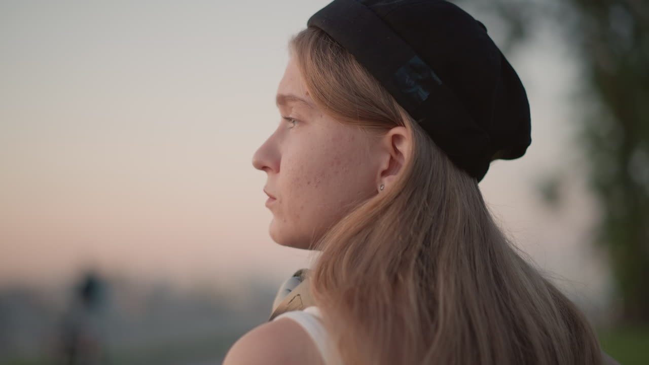 Uno sguardo allo skyline serale di un escursionista caucasico che legge, poi alza lo sguardo di profilo, cuffie intorno al collo, stato d'animo contemplativo, orizzonte distante, cielo dai colori pastello tenui, tranquilla pausa di riflessione