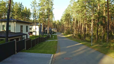 Luftdrohnenaufnahme-Beim-Überfliegen-Der-Straße-Mit-Reihen-Von-Bunglows-Auf-Der-Einen-Seite-Und-Grünen-Waldbäumen-Auf-Der-Anderen-Seite-Während-Der-Abendzeit