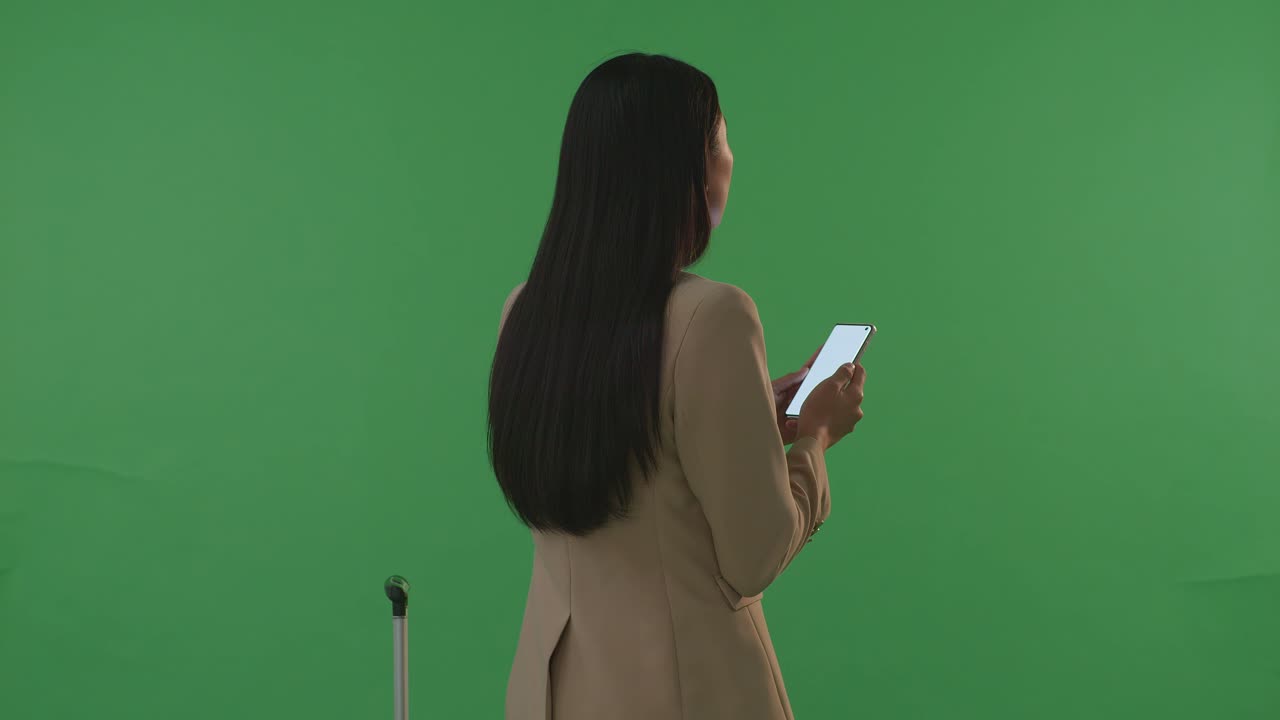 뒷모습 아시아 사업가 여성 여행자 짐과 함께 스마트폰을 사용하면서 녹색 화면 배경 스튜디오에 서서, 해가 지는 시간