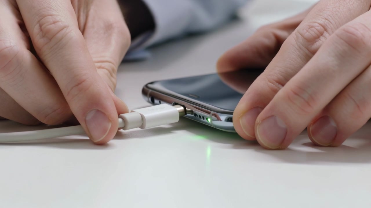 Uomo che si allunga in camicia azzurra che inserisce un cavo di ricarica bianco nella porta del telefono, con LED verde acceso