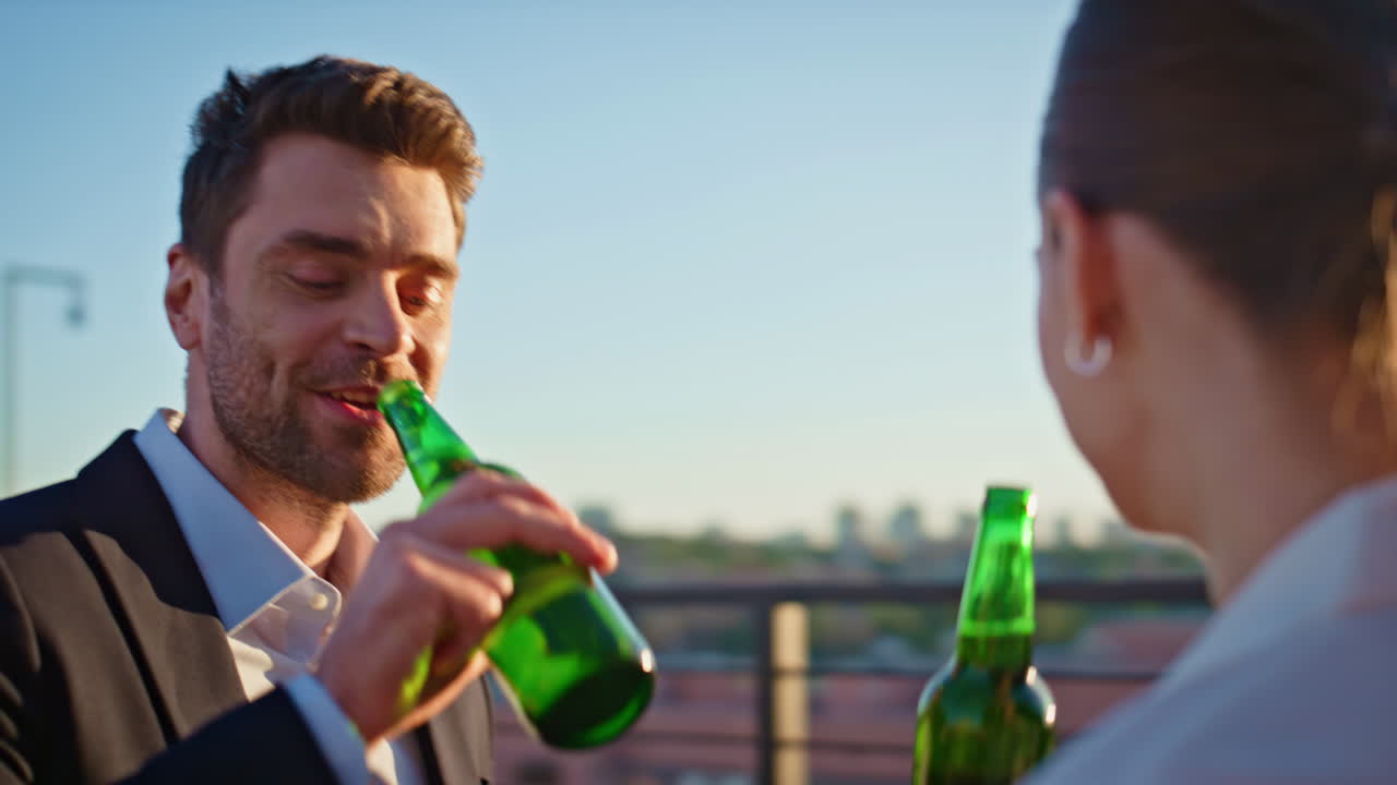Laughing coworkers clinking beer bottles spending sunset at rooftop closeup