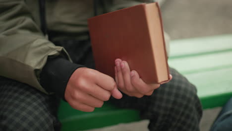 Close up side view of person in green jacket and checkered pants holding brown cover novel in right hand while seated on green bench with partial view of backpack nearby outdoors