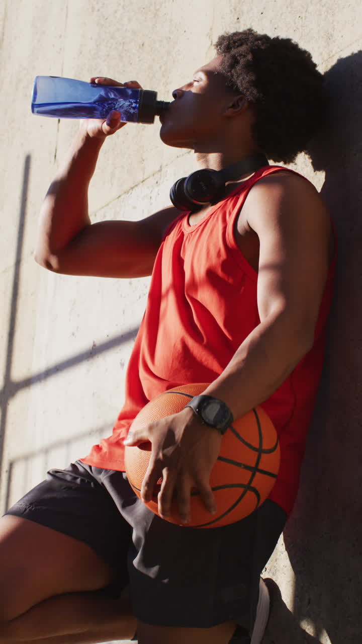 disparo vertical de un hombre biracial apoyado en la pared, bebiendo agua, sosteniendo una pelota de baloncesto
