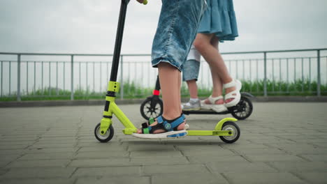 leg view of a child riding a green scooter beside their mother, who is carrying the younger child on a scooter