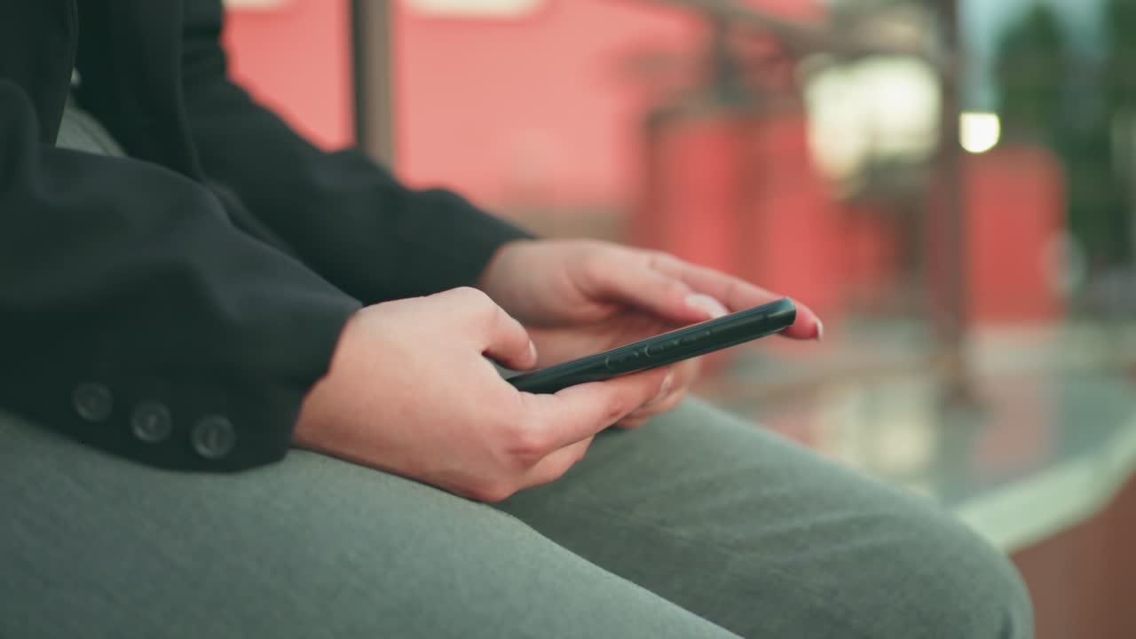 Partial view of lady with artificial nails using phone in both hands while seated, wearing black blazer and grey pants, with soft bokeh background of lights and urban structures