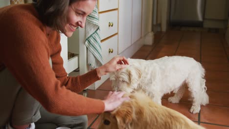 Lächelnde-Kaukasische-Frau-Streichelt-Ihre-Hunde,-Die-Zu-Hause-In-Der-Küche-Aus-Einer-Schüssel-Essen