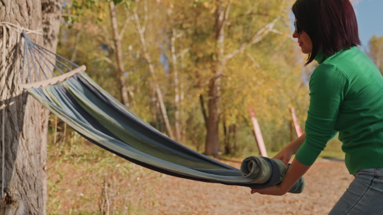 Woman setting hammock by trees preparing campsite, unrolling fabric, securing strap and tying knot, green sweater and sunglasses, autumn foliage and soft sunlight, hands arranging rope and anchor