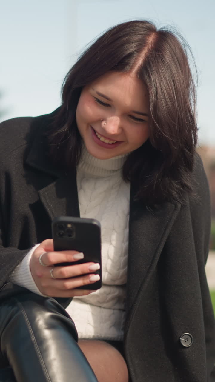 dama profesional sonriendo mientras usa su teléfono inteligente en un entorno de parque, vestida con abrigo negro y suéter blanco, disfrutando de una atmósfera al aire libre relajada con vegetación y fondo urbano borroso