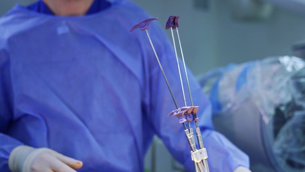 Surgeon's hands in brown latex gloves arrange long metal instruments put into patient's body. Unrecognized doctor in blue robe stands at backdrop.