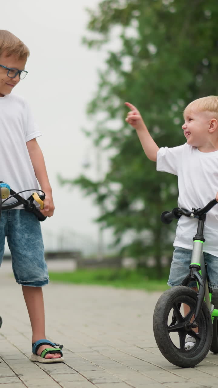 se ve a dos hermanos interactuando alegremente, con el más joven señalando a su hermano mayor mientras ambos disfrutan de estar de pie en la bicicleta, una mujer, vista desde atrás, está cerca