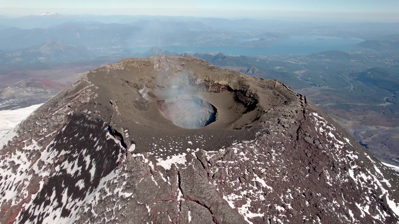 En hisnande utsikt från toppen av en vulkan avslöjar en stor krater omgiven av stenig terräng, med en fridfull sjö synlig i fjärran mot en klarblå himmel