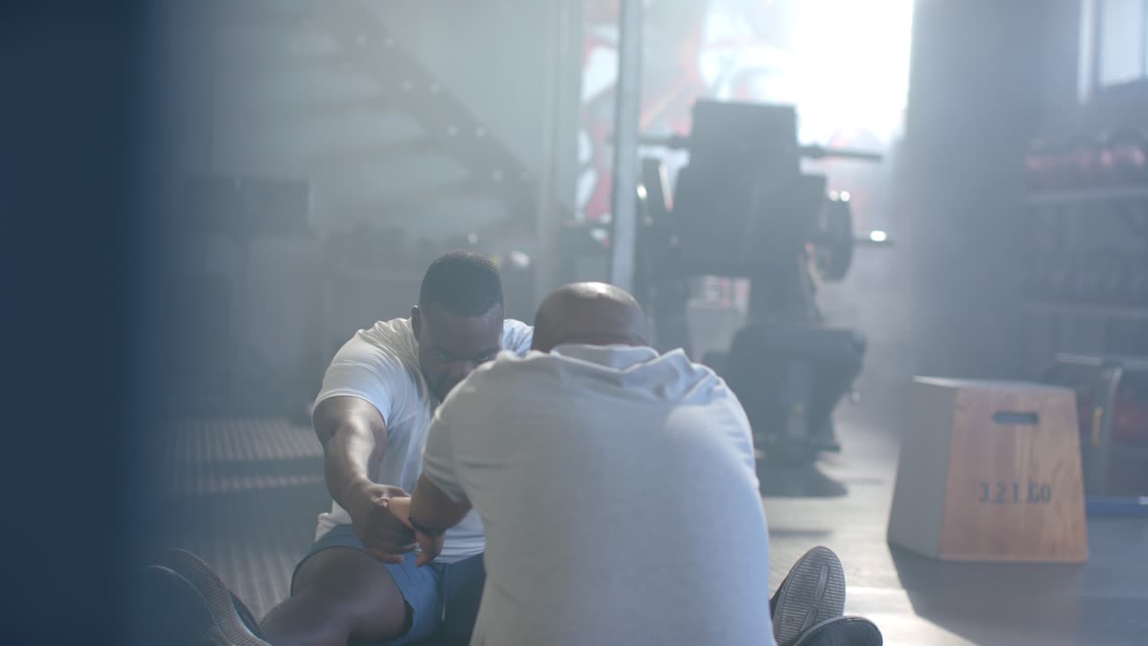 Two African men exchanging smiles and doing partner sit-ups on gym mats, pulling hands for core