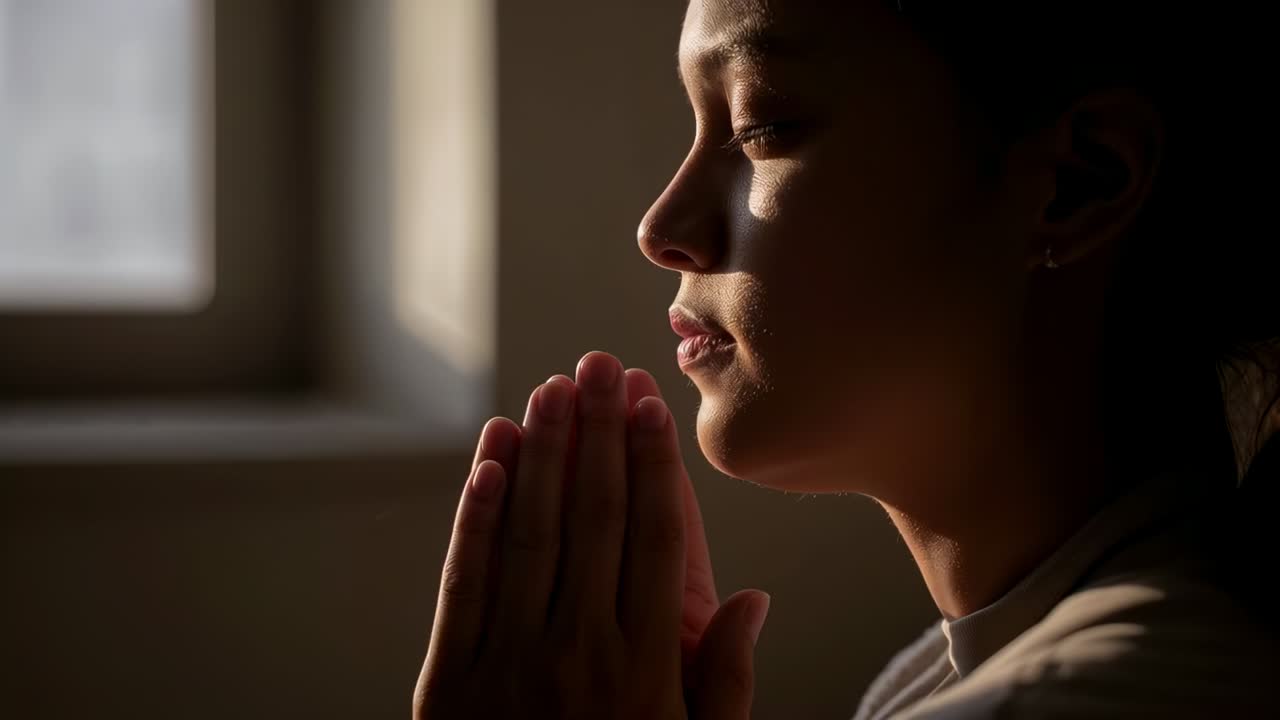 Donna in preghiera con le mani giunte vicino al mento, illuminata dalla luce della finestra, che respira lentamente in silenziosa riflessione