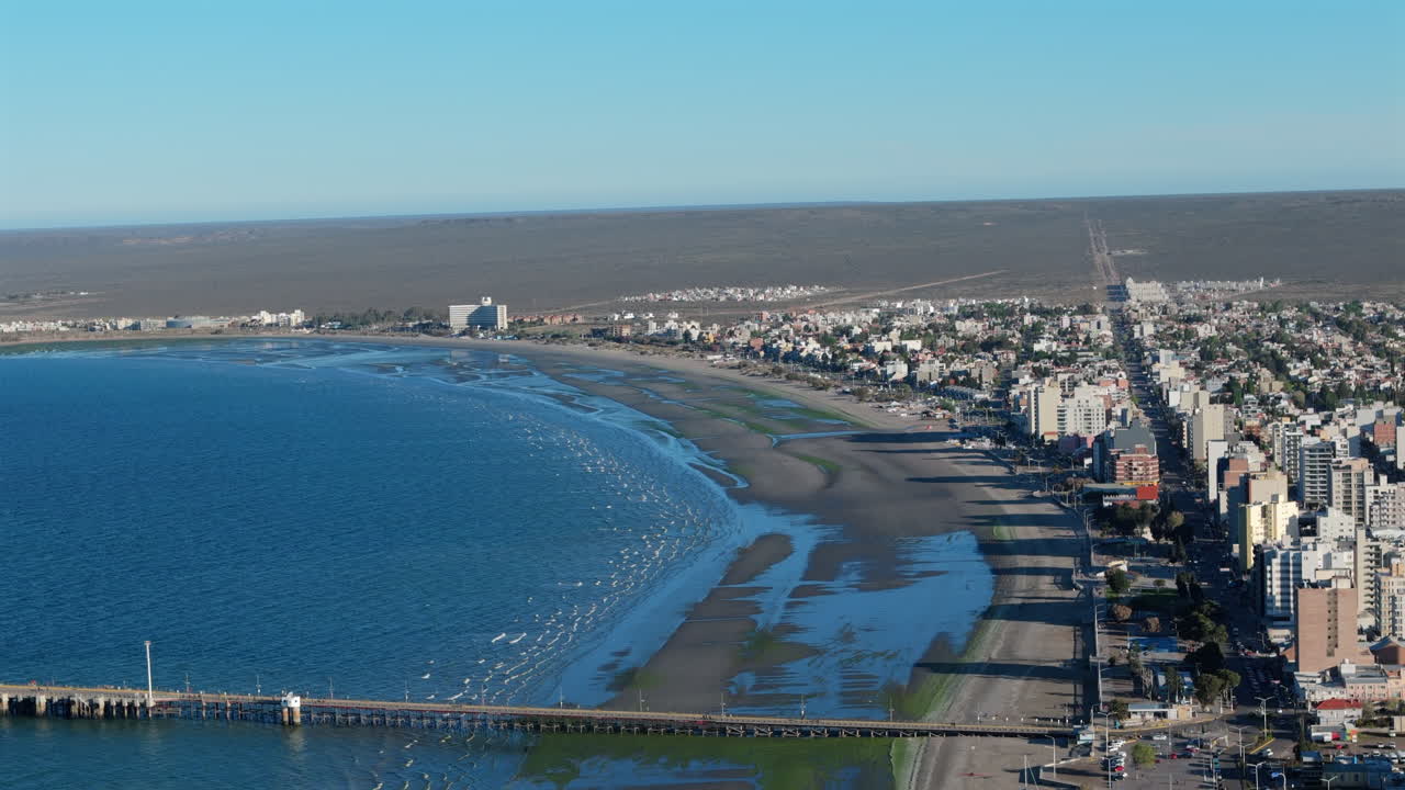 4K 60fps drone shot over Puerto Madryn’s coastline. Dock and building shadows cast on the shoreline under a clear blue sky. Distant horizon.
